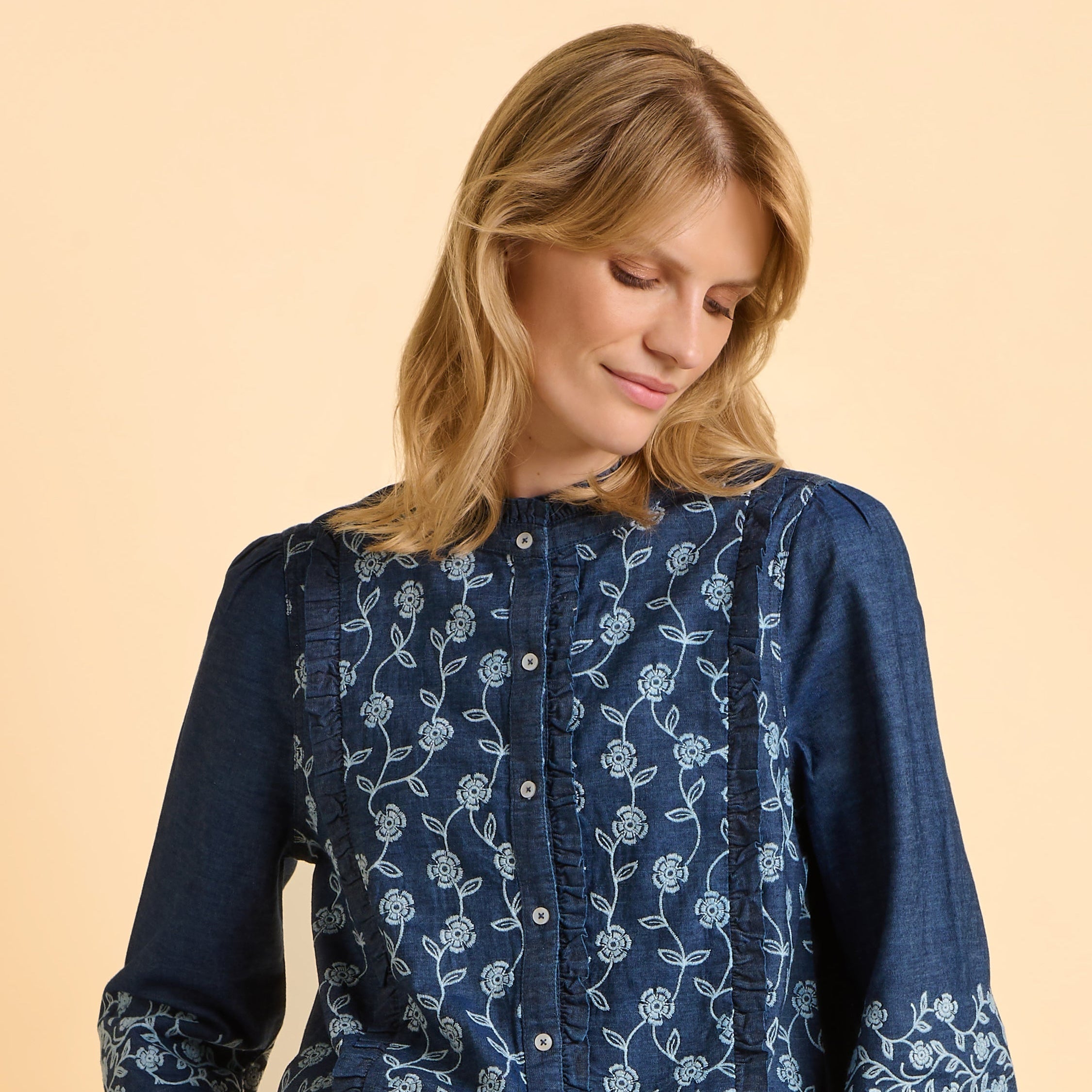 Female model wearing a blue floral embroidered denim blouse against a beige background