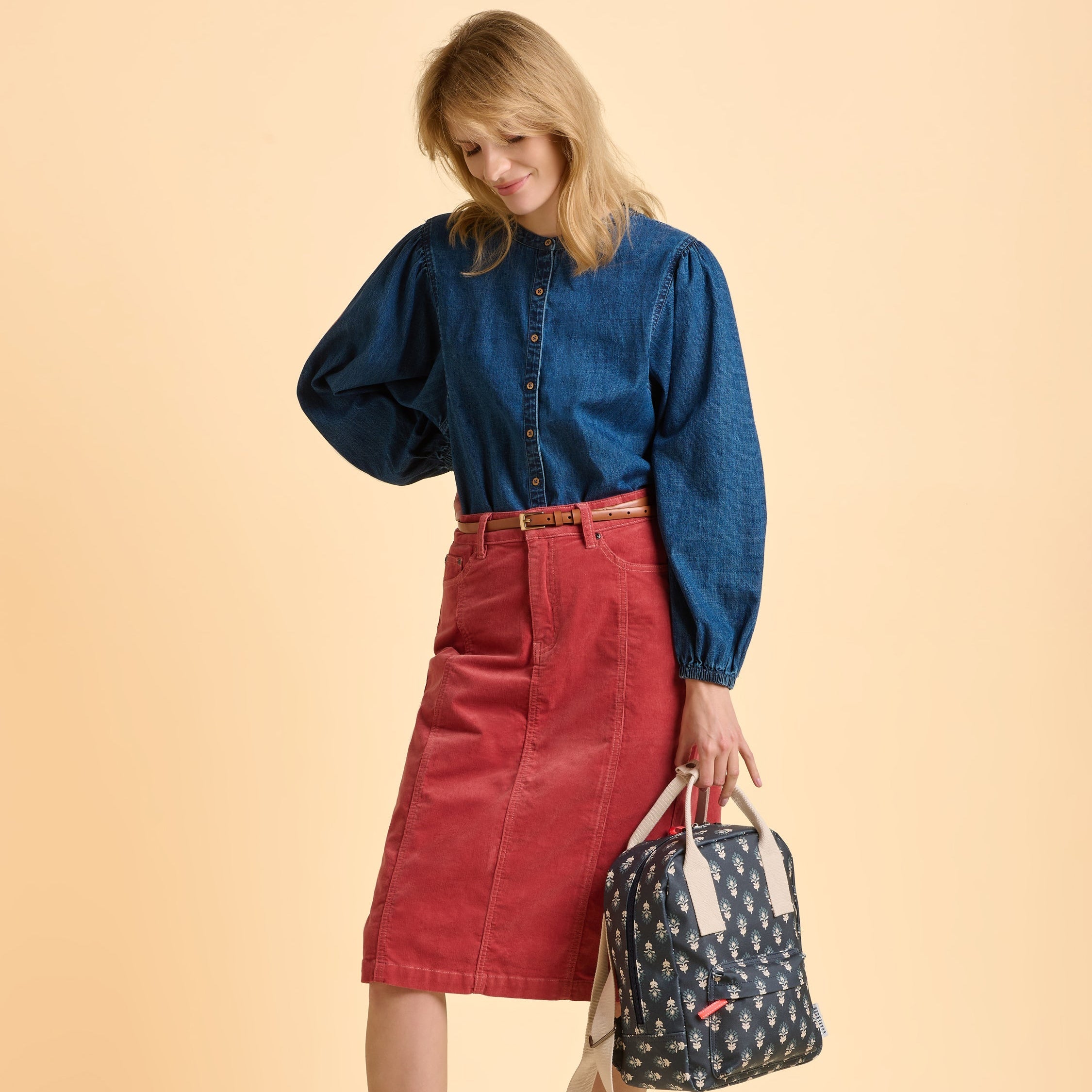 Female model wearing a blue denim shirt and red skirt holding a patterned bag on a beige background