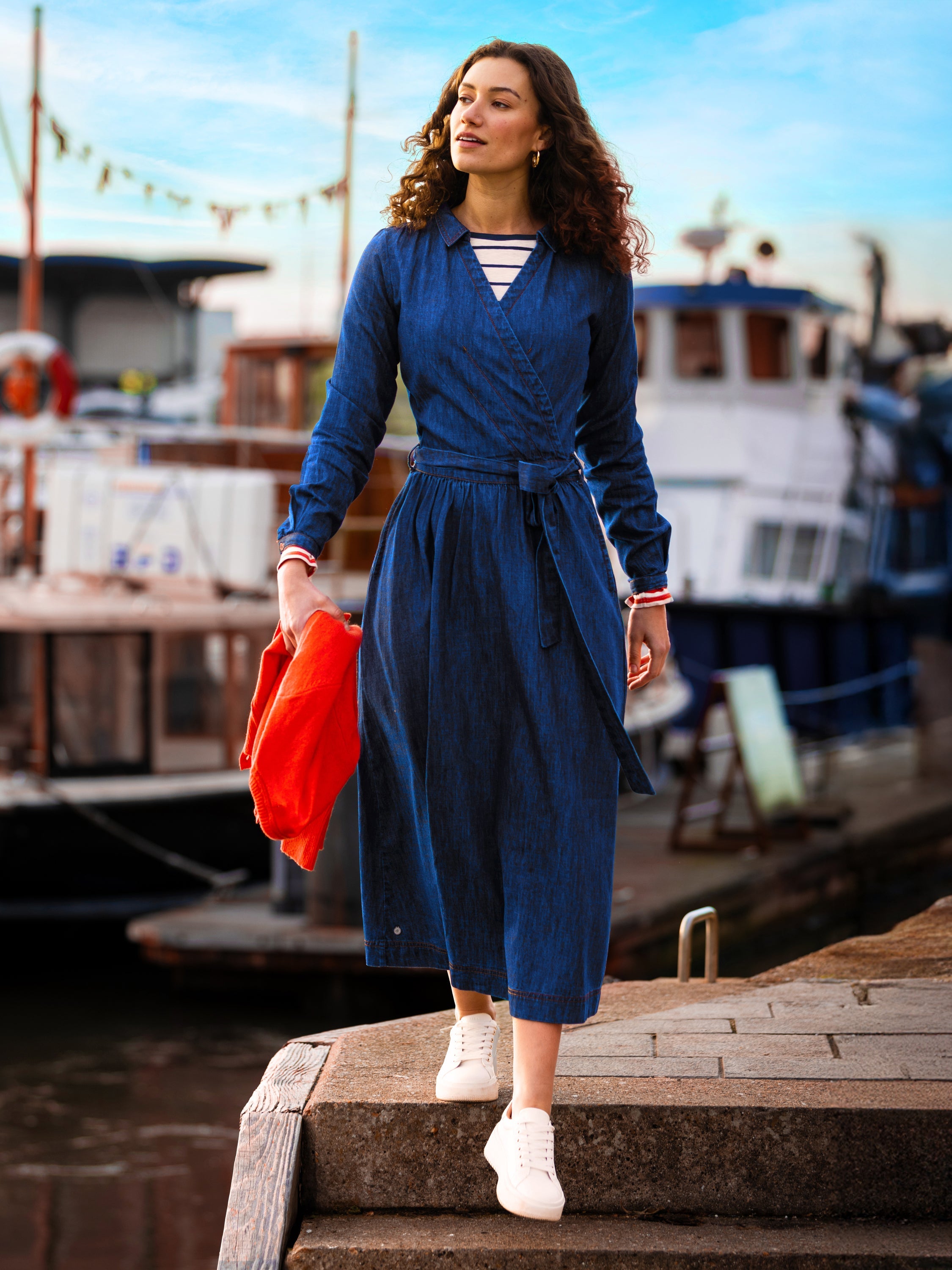 Female model wearing a blue denim wrap around dress by a boat port.
