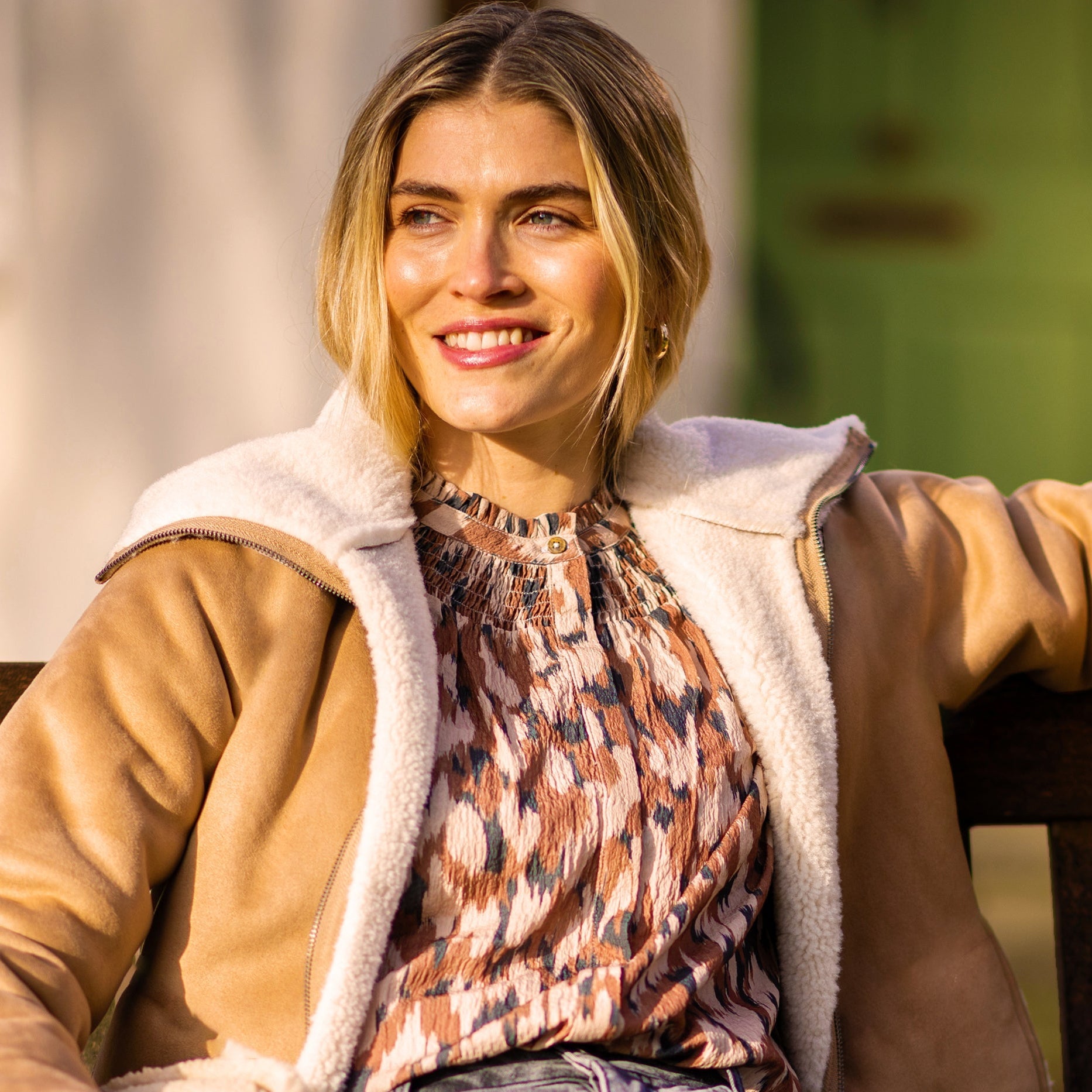 Woman sitting on a bench wearing a brown coat with white fur lining, patterned shirt, and blue jeans.