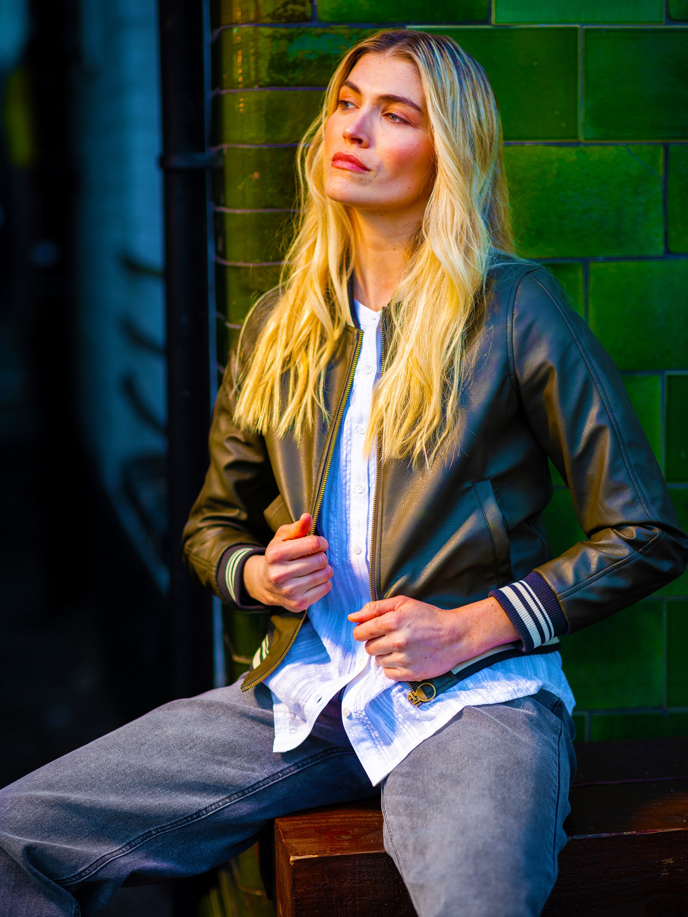 Female model wearing a leather jacket from the Brakeburn womens coat collection, sat on a bench by a city wall.