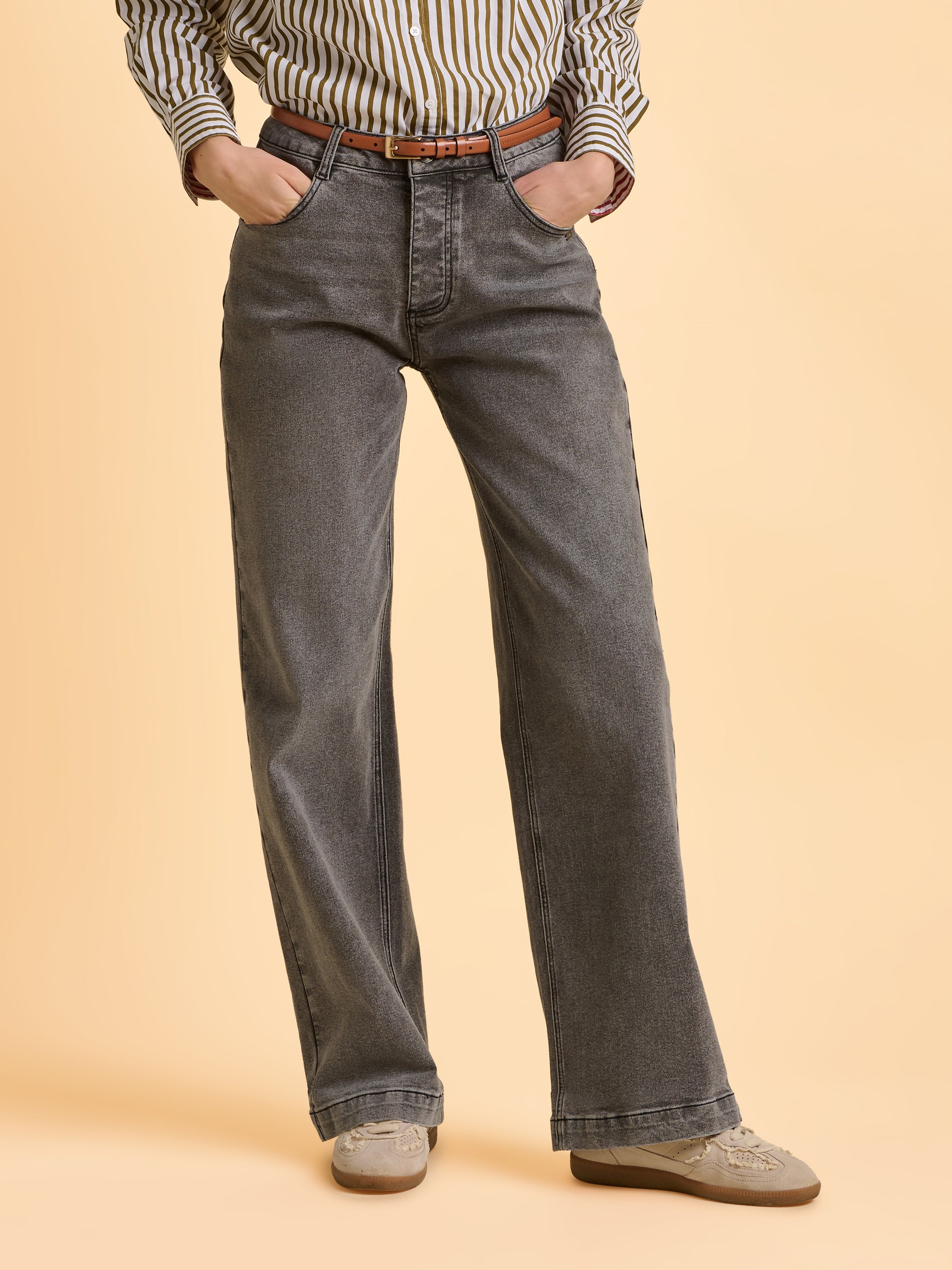 Female model wearing grey Brakeburn jeans with a brown belt, stood in a studio setting posing for the camera