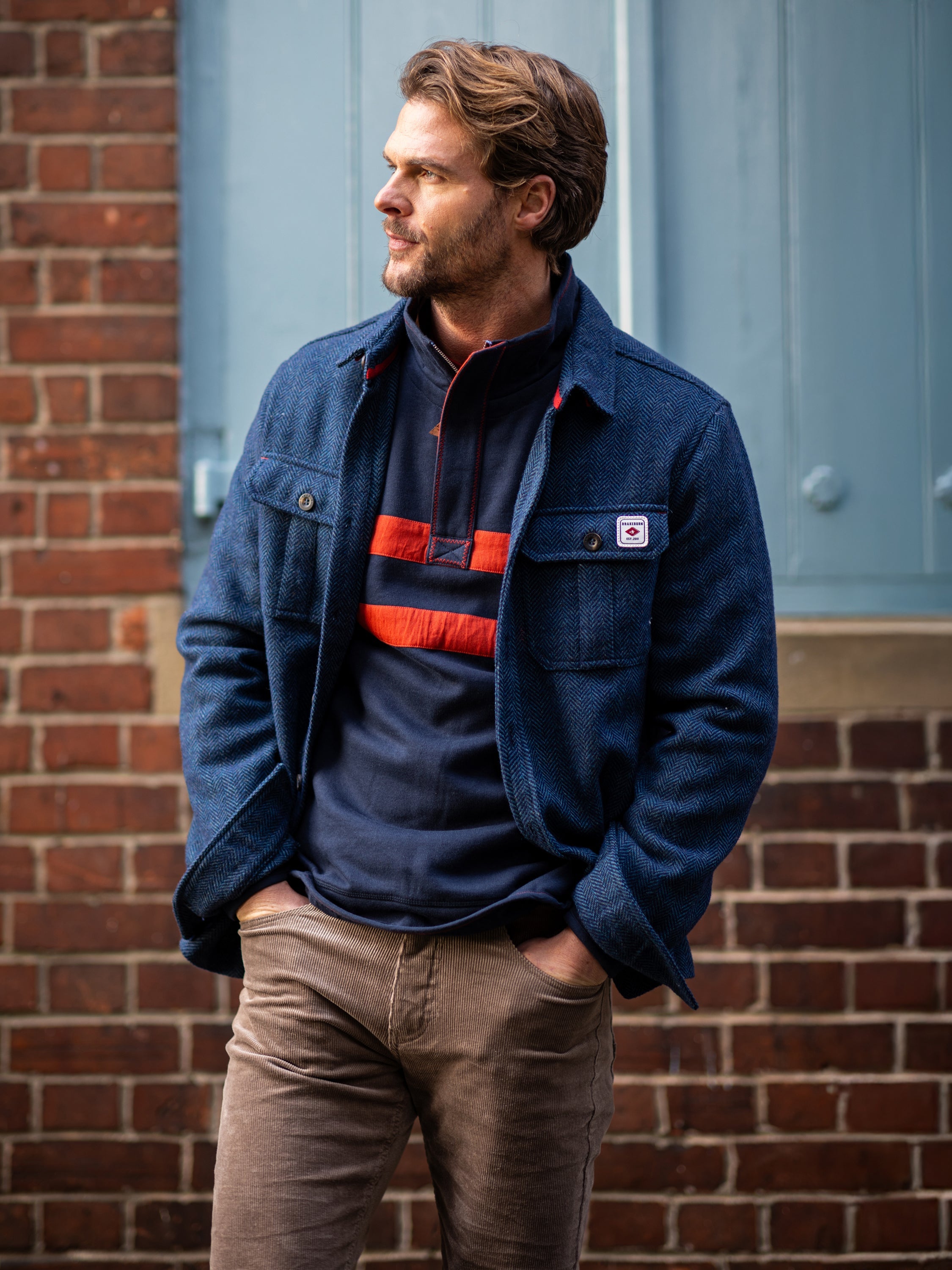Male model wearing a Brakeburn blue jacket and Brakeburn blue jumper with corduroy trousers in front of a brick wall