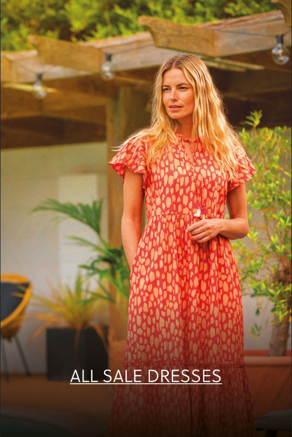 Woman in a red patterned dress standing near a sunny patio garden – featured in the All Sale Dresses section