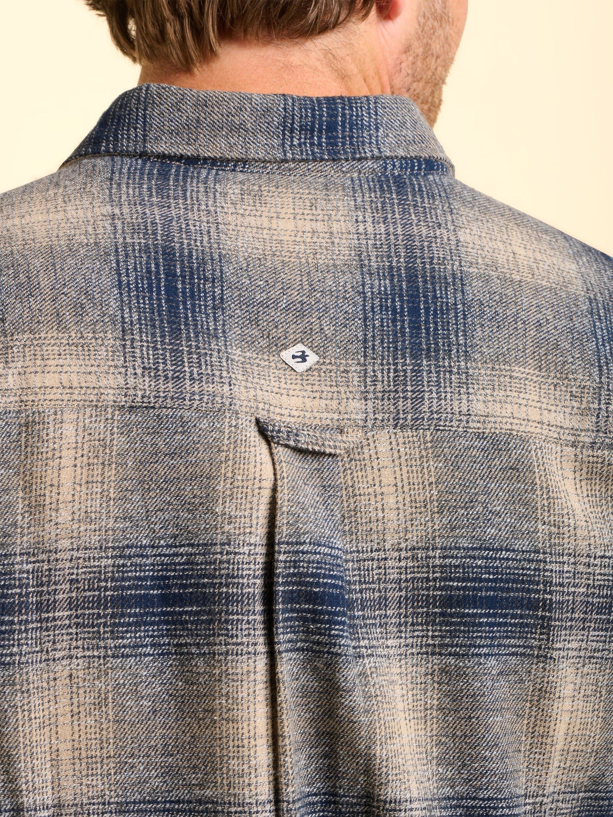 Close-up of the back of a man wearing a blue and grey plaid check shirt with a Brakeburn logo in the centre.