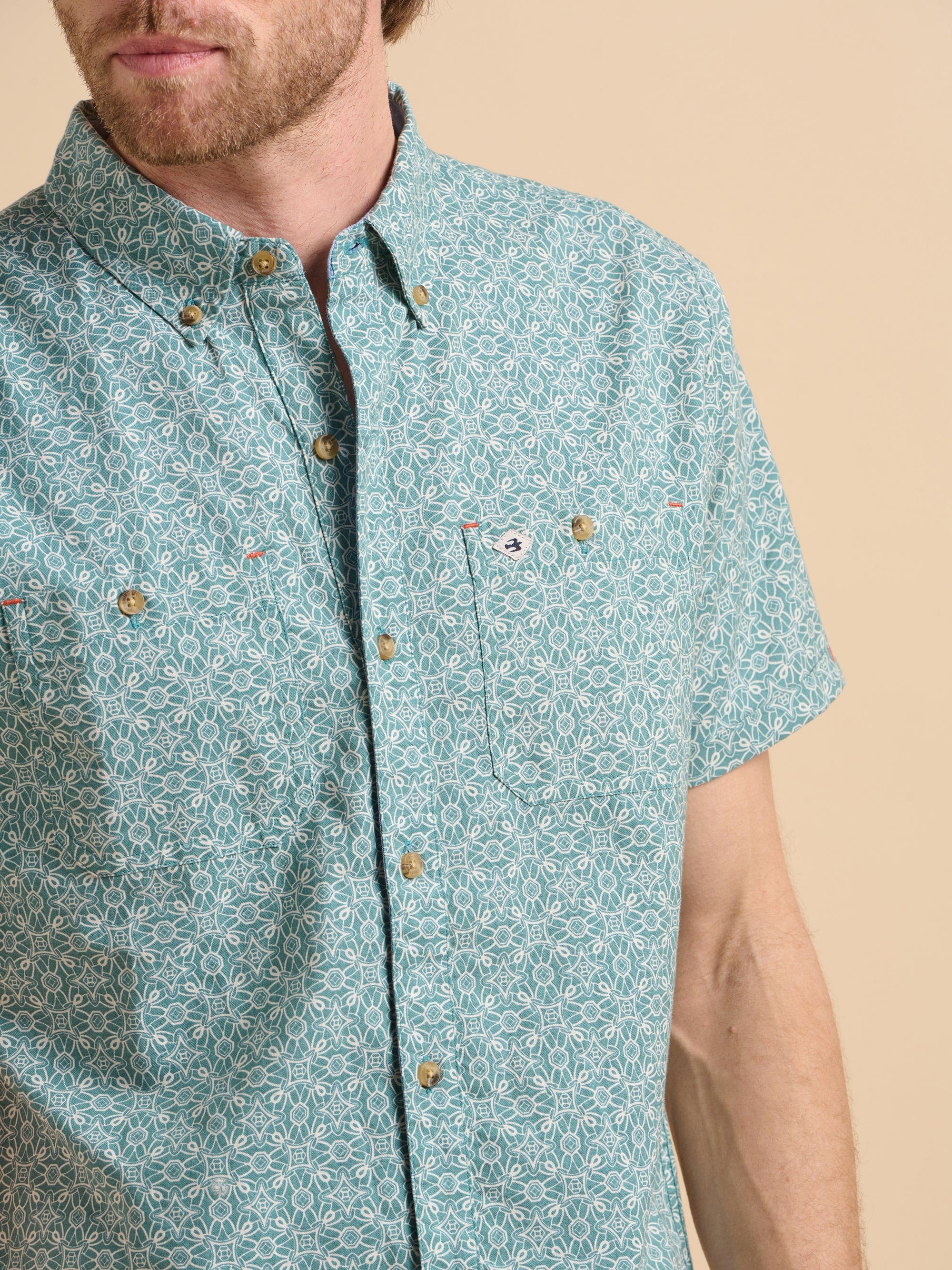 Man wearing a teal patterned short sleeve shirt standing in front of a beige background.