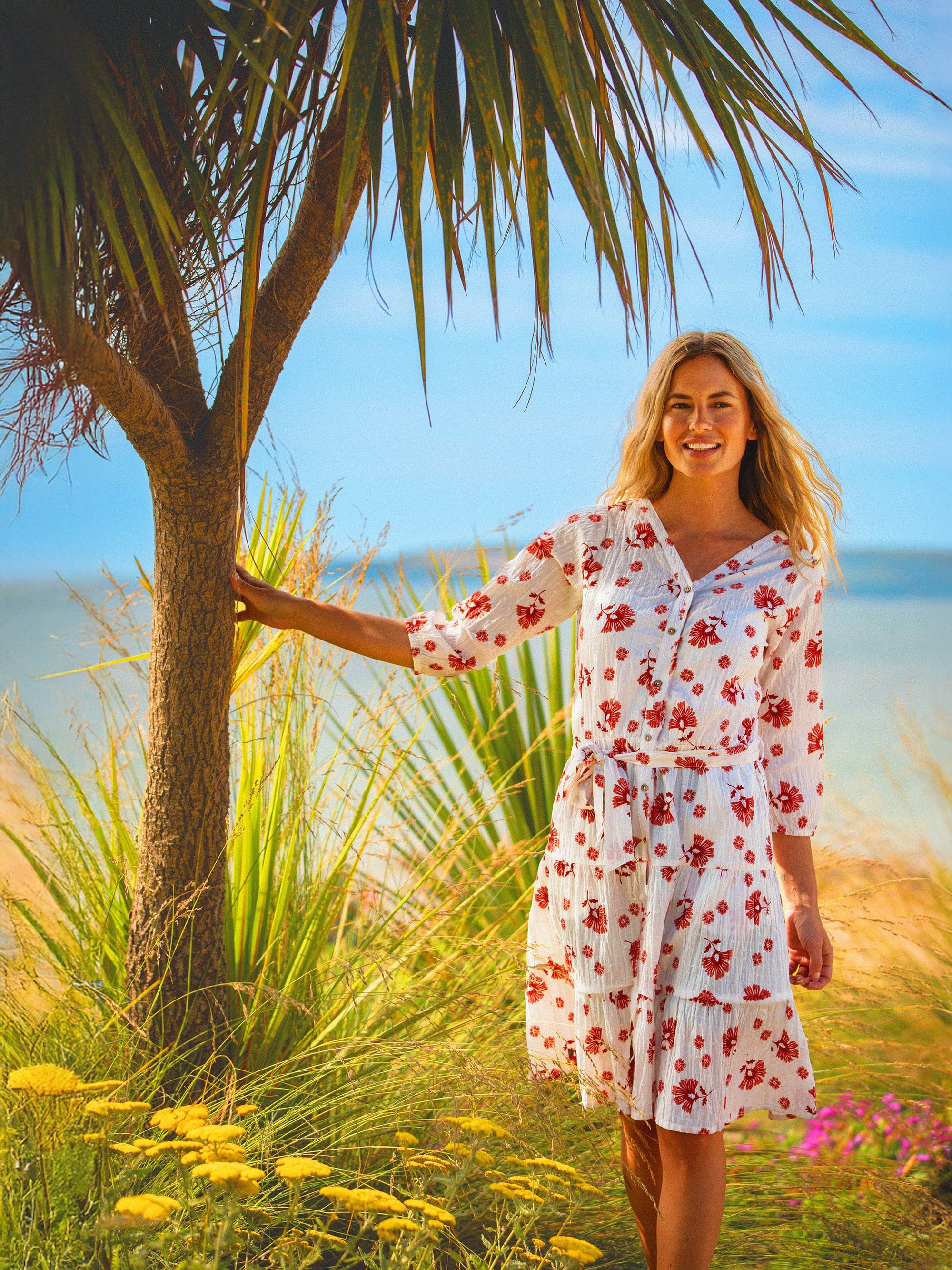 A woman standing next to a palm tree wearing a knee-length white and red floral dress with 3/4 sleeves and a tie waist.