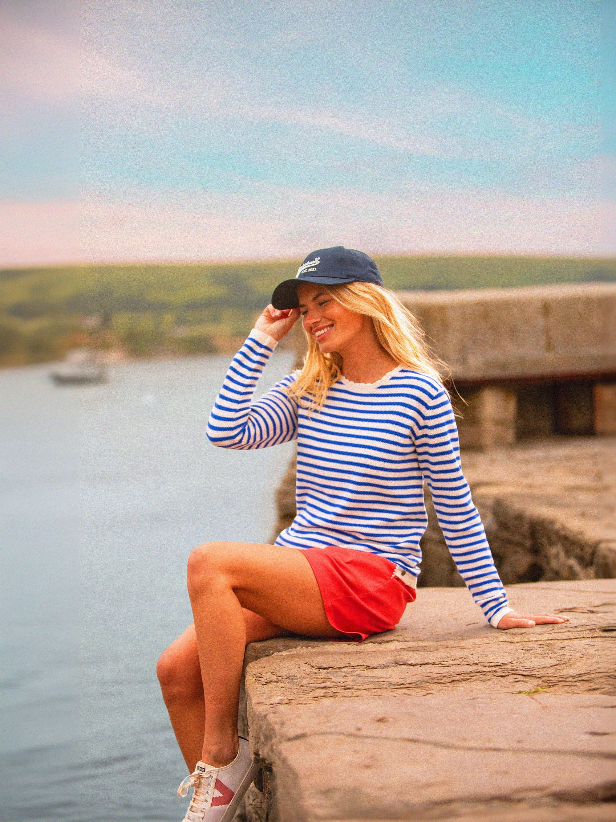 Woman in a striped knitted jumper and red shorts sitting by a lake with a scenic background.